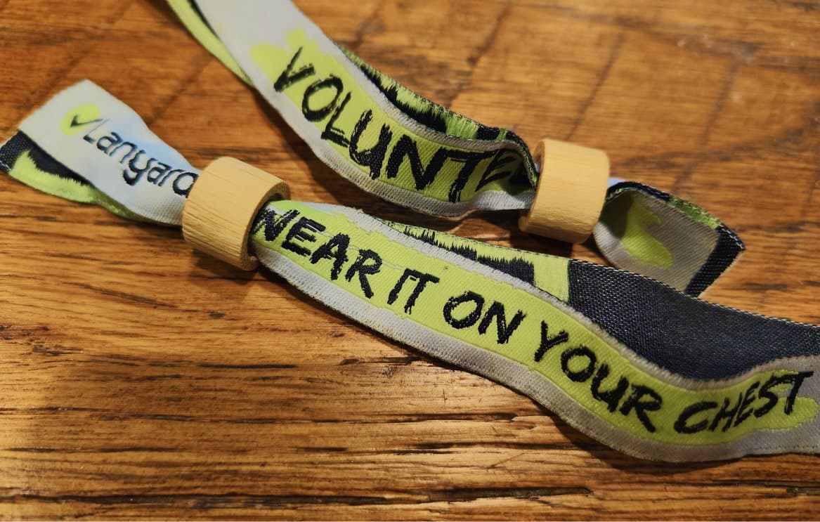 Two woven Lanyard wristbands on a wooden table, printed 'VOLUNTEER' and 'WEAR IT ON YOUR CHEST'.